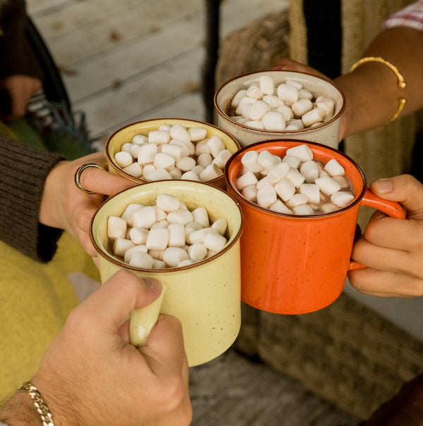 Four mugs filled with hot chocolate and marshmallows being held by people outdoors.